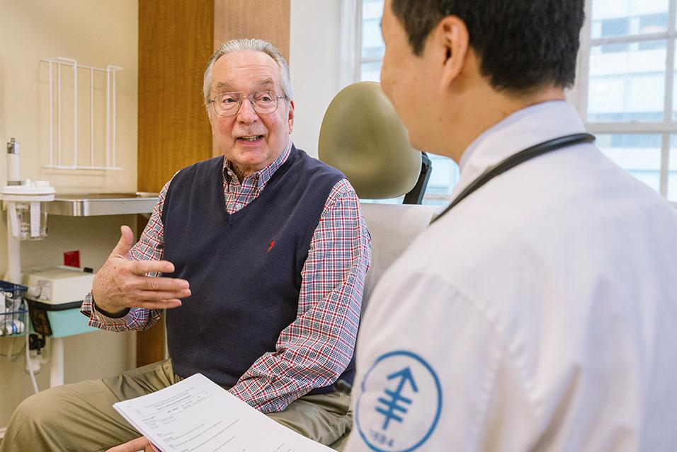 An elderly man sits in an exam chair and is speaking to an MSK clinician whose back is to the camera. 