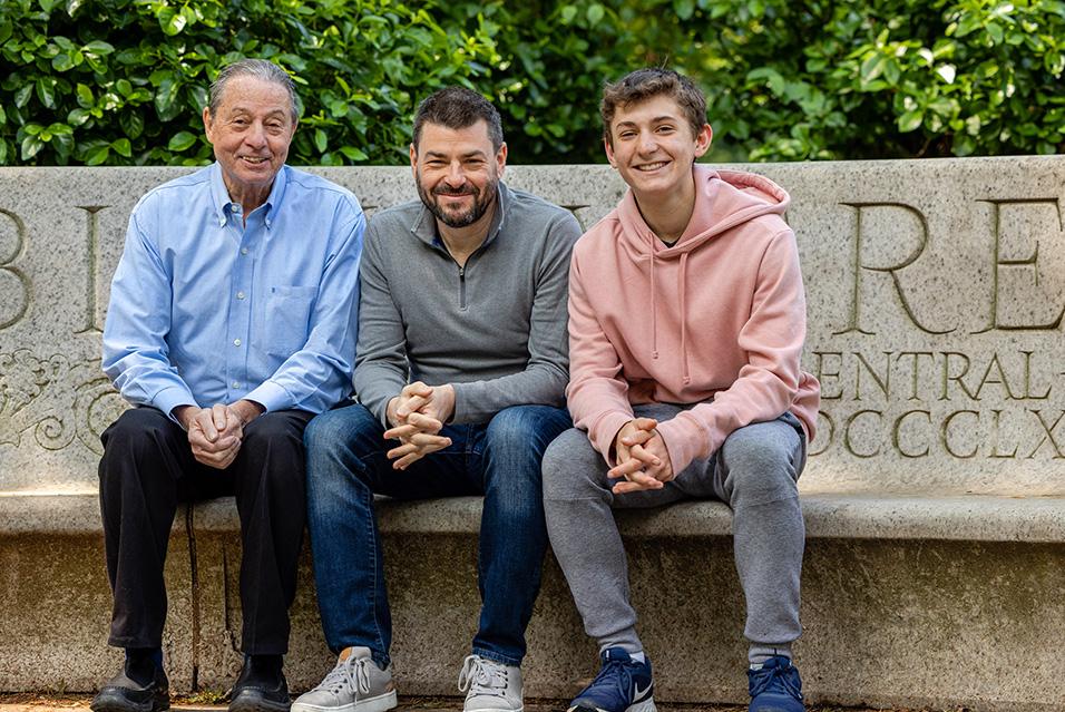 Three people sit, smiling, on a marble bench, with greenery behind them. From left: an older man, a middle-aged man, and a teenage boy. 