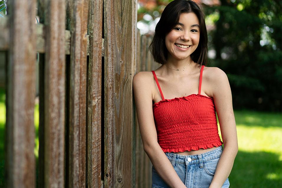 A young woman wearing a red top and jeans is standing outside, leaning against a fence and smiling. 